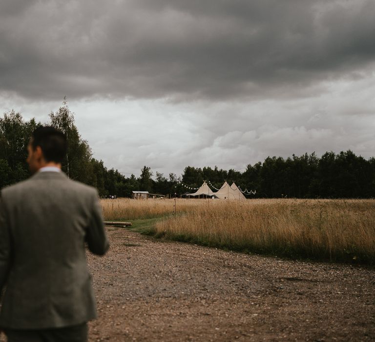 Bride in white lace open back wedding dress and flowers inter hair holds hands with groom in grey herringbone wedding suit walk towards wedding tipi at Wellington Wood Norfolk for late summer wedding