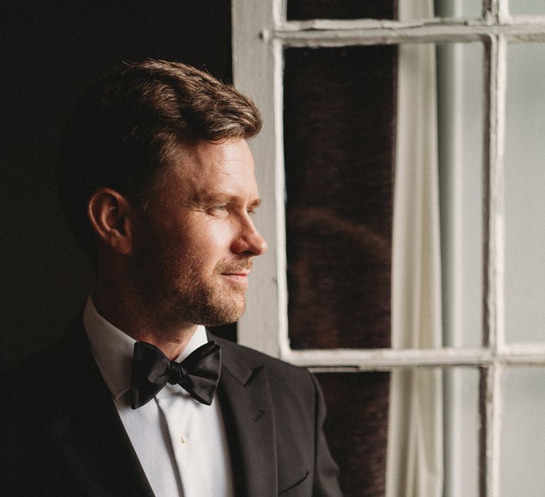 Groom in a black suit and bow tie looking out the window 