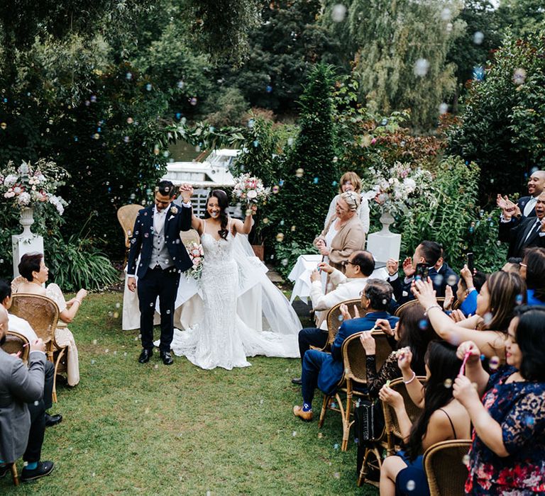 South Asian couple throw hands in the air with joy as they stand in stunning fitted garments 