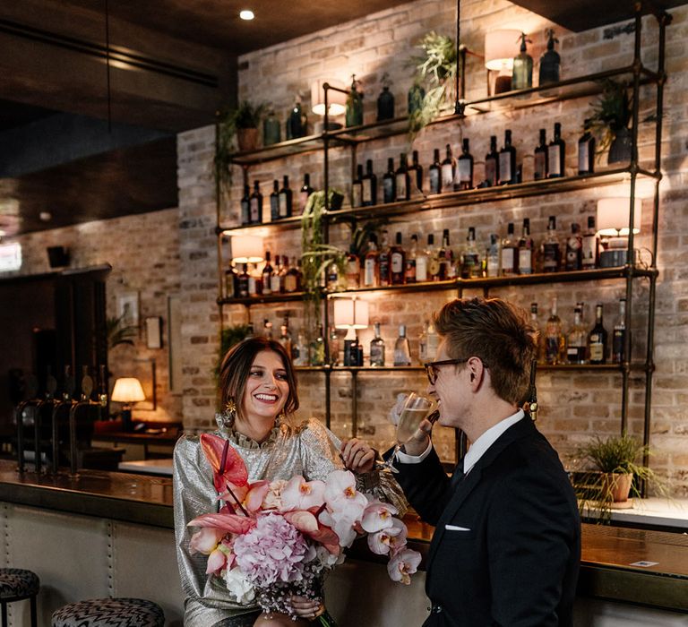 Bride in short shaped high neck short gold wedding dress & Groom wears black slim fit suit raising a glass in modern London bar 