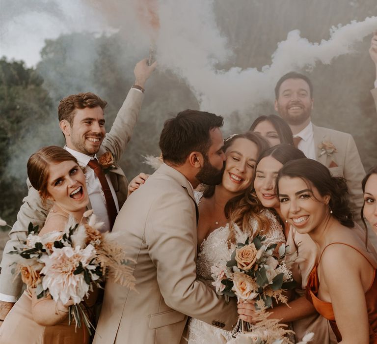 Groom in beige suit kisses cheek of bride in homemade lace wedding dress whilst bridesmaids gather around them and groomsmen smile holding orange and white smoke bombs at outdoor wedding in Bedfordshire