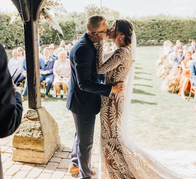 Groom in navy suit and tanned brogues kisses Bride in Rue De Seine (wedding dress) as wedding guests cheer at Barn wedding 