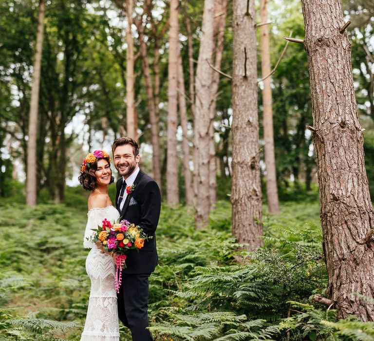 Smiling bride in off the shoulder Grace Loves Lace wedding dress and flower crown holding colourful bridal bouquet stands with smiling groom in black suit in woodland after outdoor wedding ceremony at The Bridal Barn in Claverley