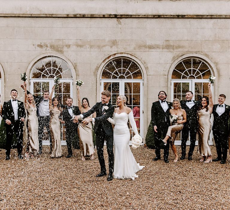 Wedding party portrait with the brides and groom popping champagne. The groomsmen wear tuxedos and the bridal party champagne gold bridesmaid dresses 