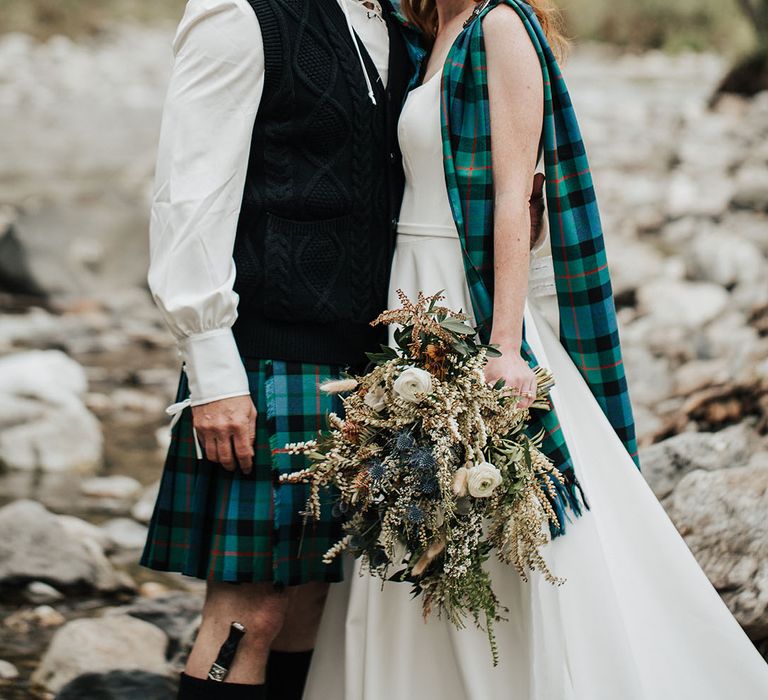 Bride and groom in a tartan kilt and sash standing in a stream holding a boho wedding bouquet with anemones 