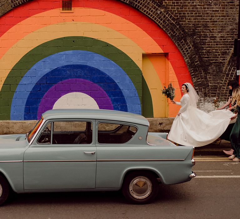 Bride's dress is held up by bridesmaids as she walks in front of painted rainbow backdrop and next to vintage blue car