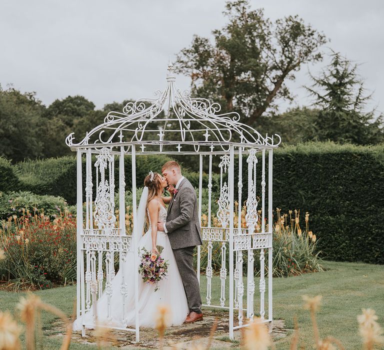 Bride and groom share a kiss in white iron birdcage at rustic country house wedding venue 