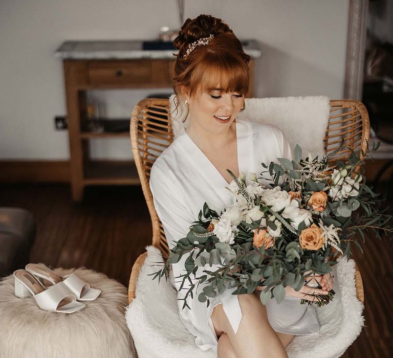 Bride in white satin dressing gown and fluffy socks with satin white mules on fluffy stool with wedding bouquet