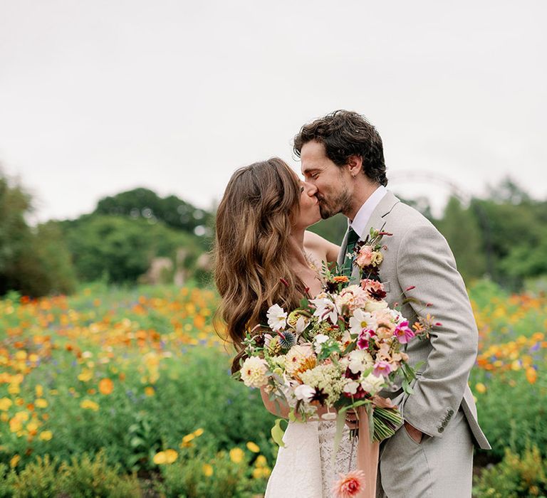 Bride and groom share a kiss in a field of wildflowers as the bride holds a wildflower wedding bouquet 