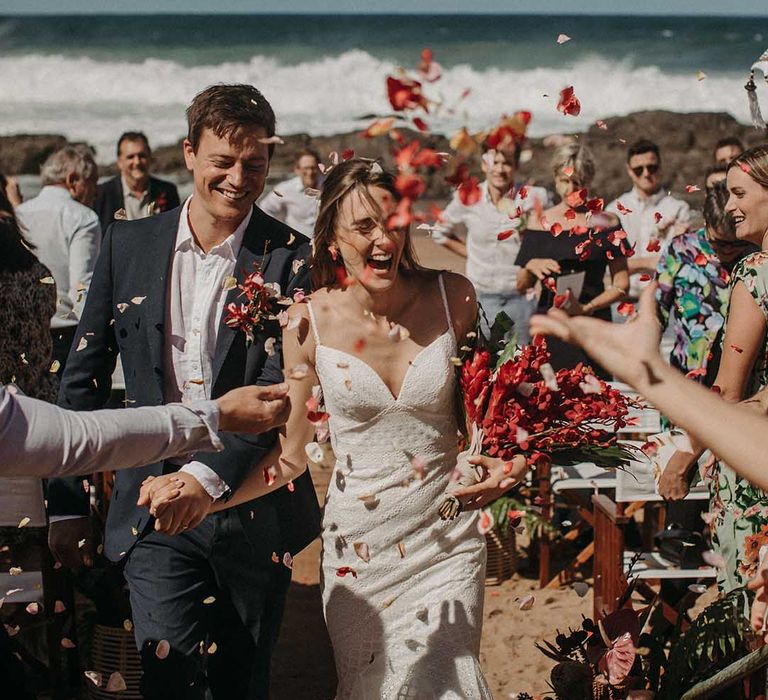 Bride in v-neck fitted lace beach wedding dress and groom in suit jacket walk through red confetti together after beach ceremony 