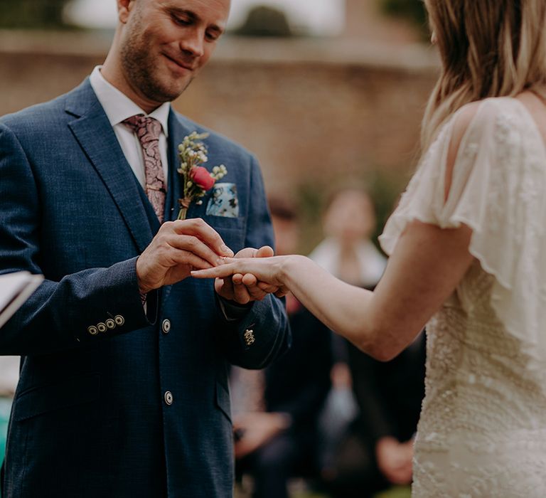 Groom in blue suit and paisley patterned tie places the ring on the bride's finger