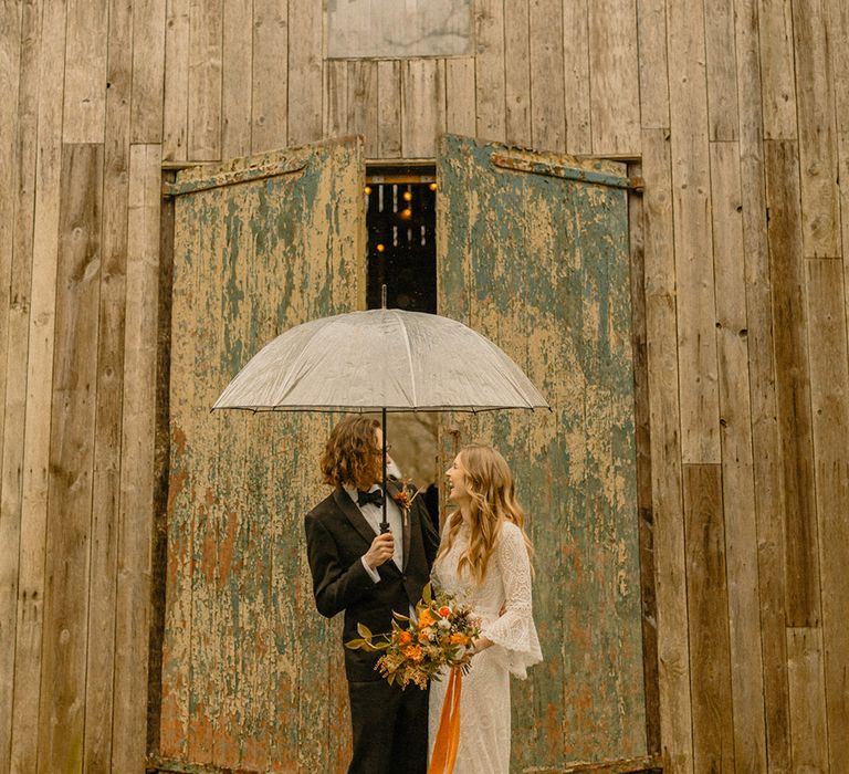 Bride in lace wedding dress with bell sleeves under clear umbrella with groom in black tie on rainy wedding day