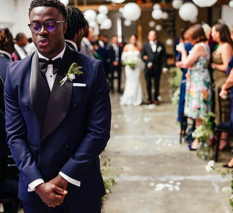 Groom in blue and black tuxedo stands at the altar and takes a deep breath as he prepares to turn around and see his bride