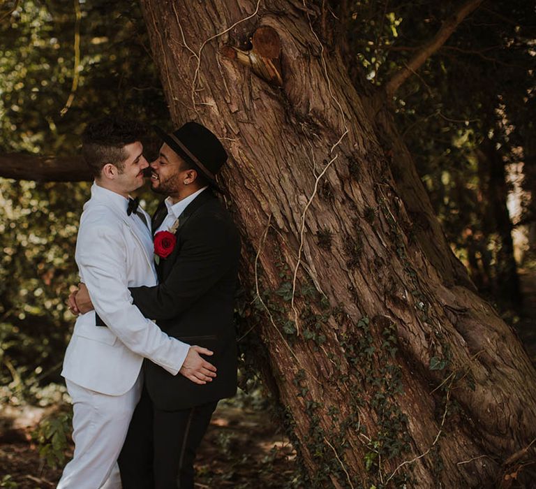 Groom in a white suit embracing his partner in a black groom suit with red Gerbra buttonhole flowers 