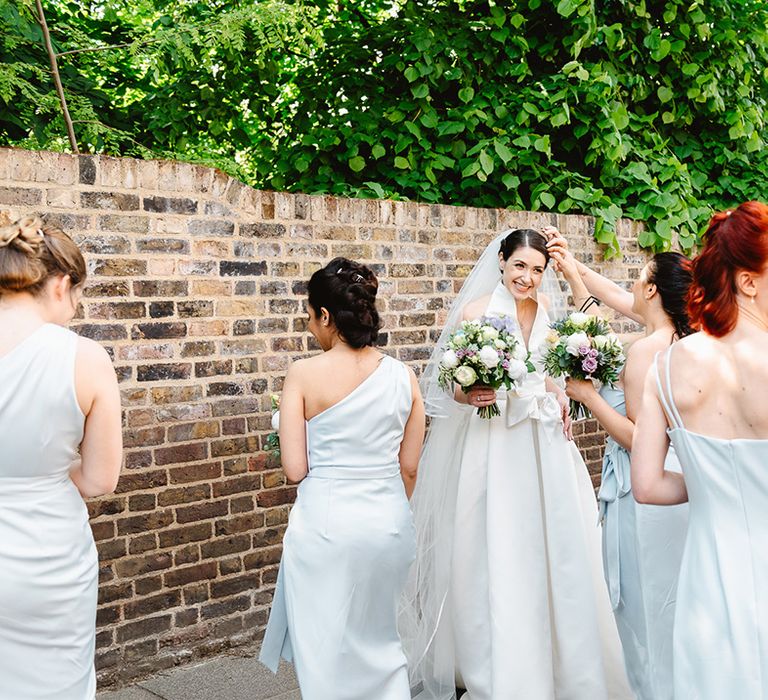 Bridesmaids in different style baby blue dresses with groom in black suit with matching baby blue tie and smiling bride as she gets help with veil