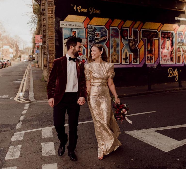 Bride in puff sleeve gold wedding dress and bow detail holds hands with groom in burgundy velvet suit jacket