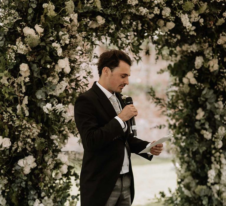 Groomsmen in morning suit and striped trousers performs a wedding reading for outdoor marquee wedding next to white flower arch