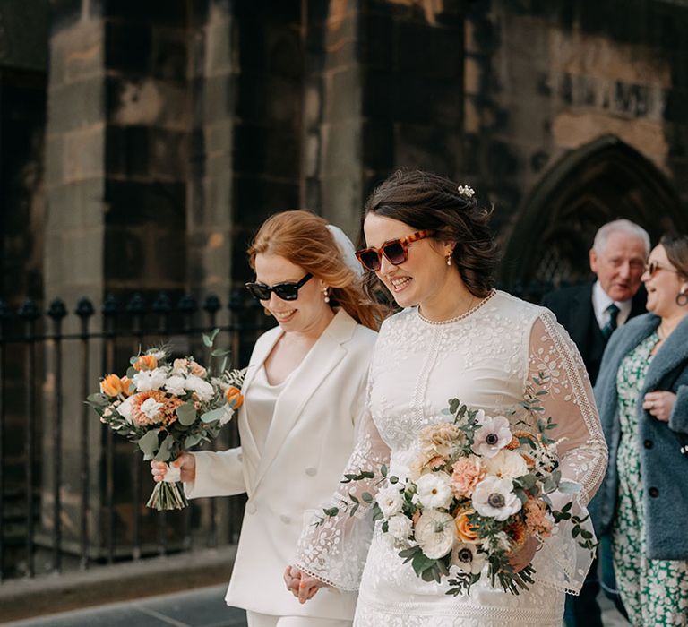 Brides walk holding hands whilst wearing sunglasses and holding colour floral bouquets 