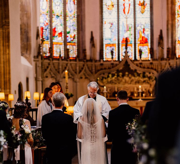 Bride in sheer back lace wedding dress stands inbetween the groom and her father as they stand at the altar for their church wedding ceremony 