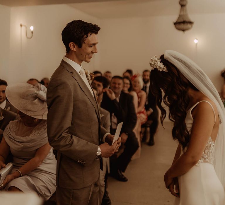 Groom smiles at the bride in neutral three piece suit as he reads out his wedding vows to the bride 