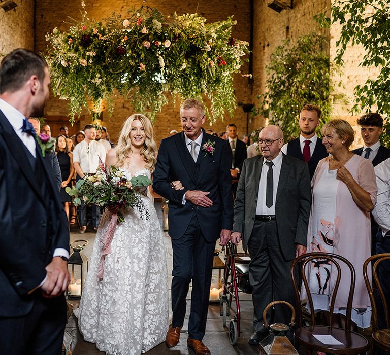 Father of the bride in blue suit and pale blue tie walks the bride down the aisle in a strapless lace wedding dress 