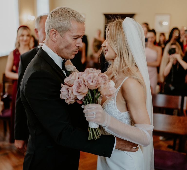 Groom in black tie embraces the bride in a beaded wedding dress with a veil and tulle gloves 