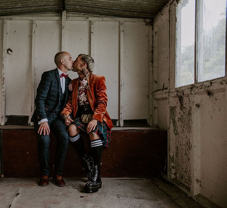 Groom wears orange velvet blazer and tartan kilt next to his groom who wears tartan suit complete with red bow tie 