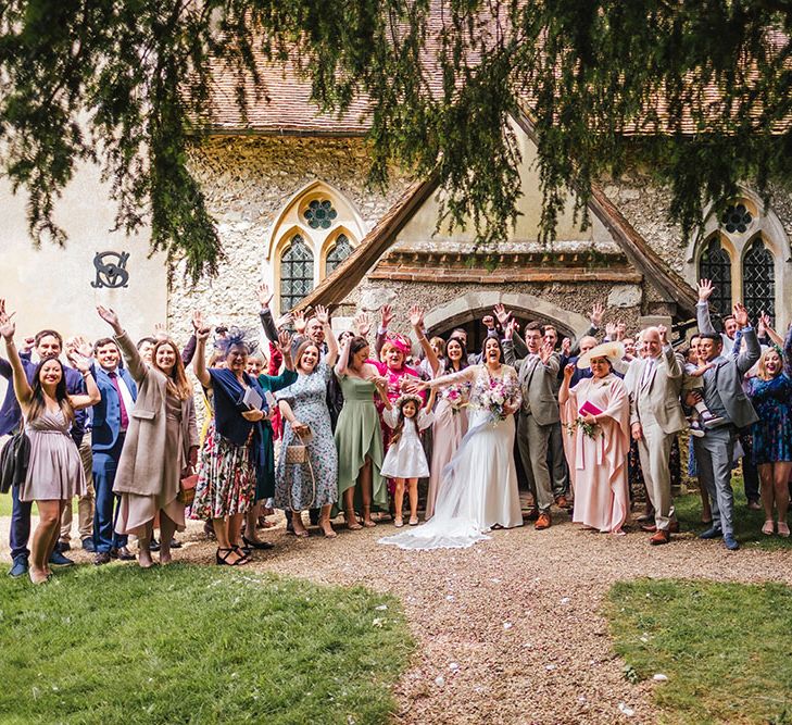 Bride & groom stand outside church on their wedding day surrounded by family and friends in colourful outfits 