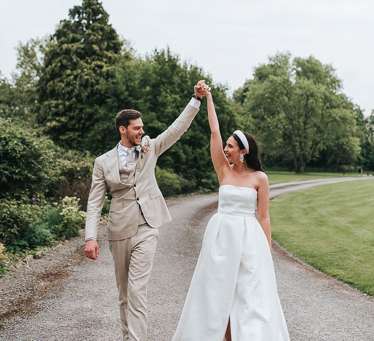 Groom in neutral three piece suit lifts hands with the bride in a brocade Jesus Peiro wedding dress and headband 