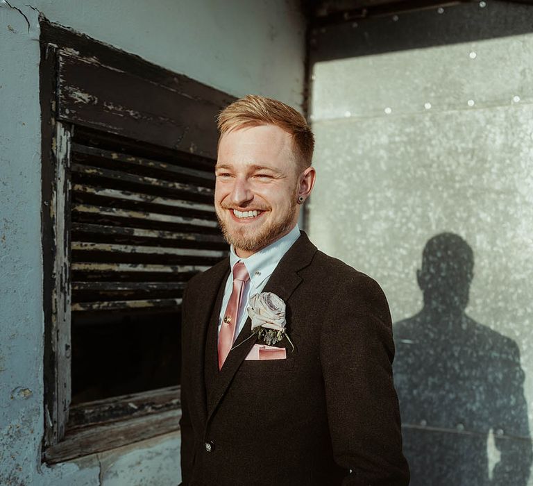 Groom in a brown suit with a pink tie and pink rose buttonhole smiling brightly 