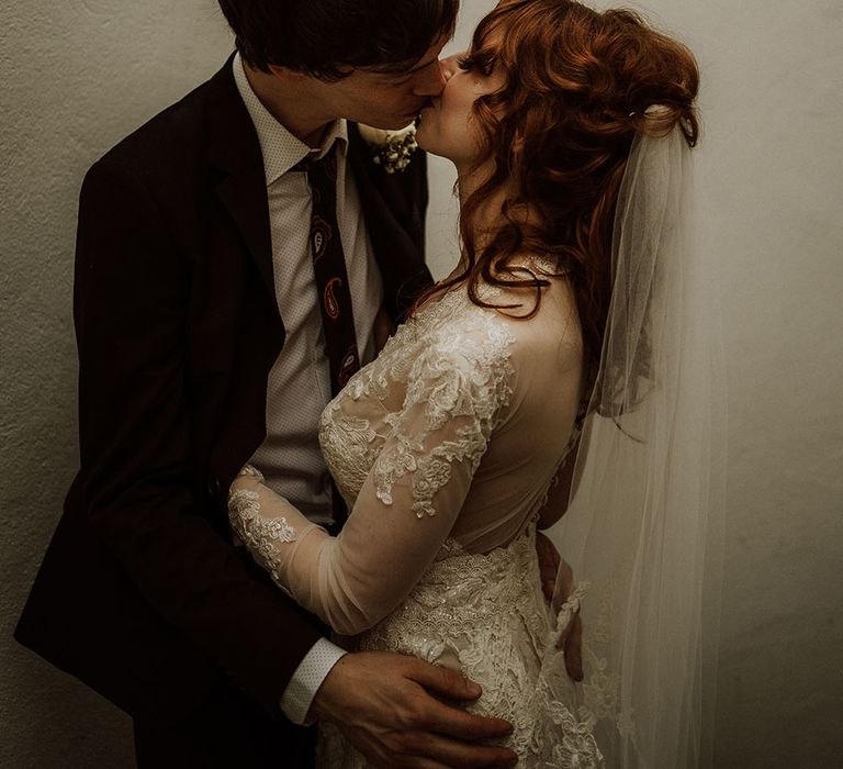 Groom wears brown suit jacket with psychedelic tie and kisses his bride wearing lace wedding dress with long sleeves 