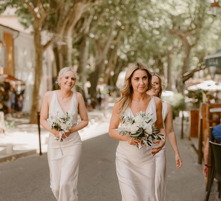 Bridesmaids wear silk white bridesmaid dresses complete with cowl necks and carry white rose floral bouquets 