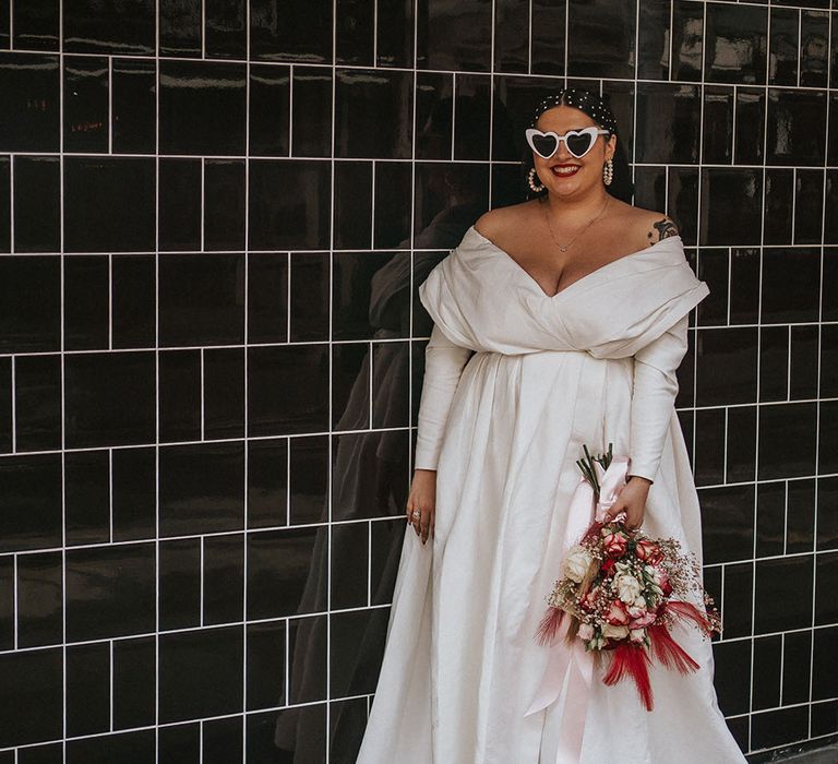 Bride wears long sleeve curve wedding dress with off-the-shoulders and plunge neckline as she holds bright floral bouquet 