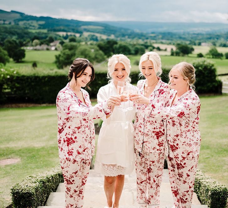 Bride & her bridesmaids in colourful pink pyjamas stand outdoors with champagne glasses on the morning of wedding day at Tithe Barn