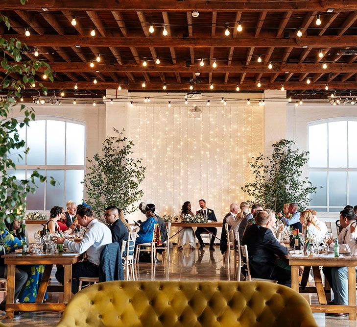 Bride & groom sit at sweetheart table in front of fairy light canopy as festoon lighting hangs from above 