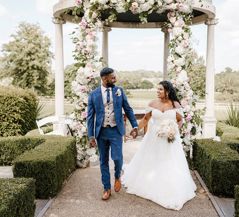 Bride holds her grooms hand who wears blue suit whilst walking away from floral archway 