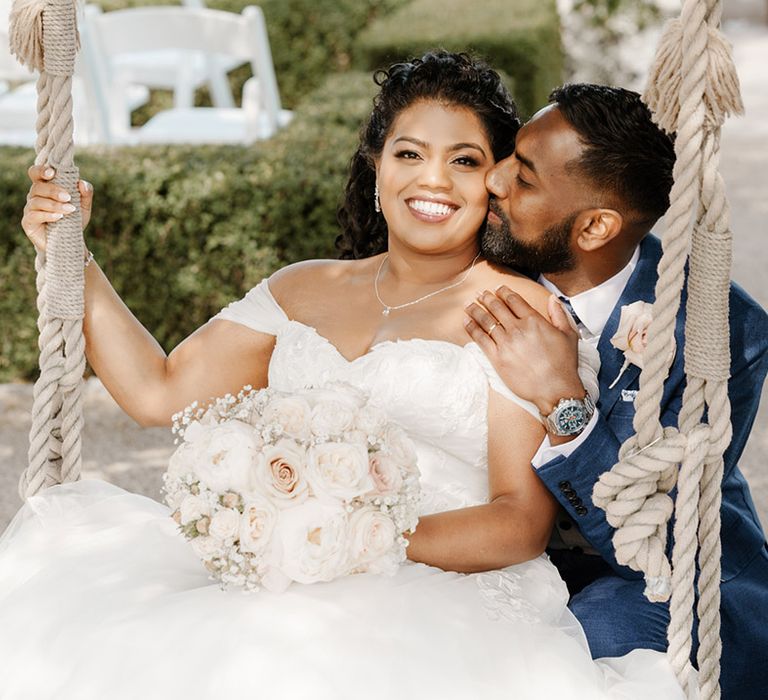 Bride sits on outdoor swing as her groom sits behind her and kisses her cheek 