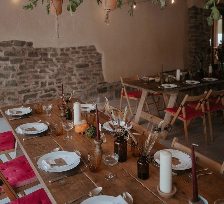 Wooden banquet tables with pumpkin decor and maroon coloured candles as terracotta pots hang above filled with plants 