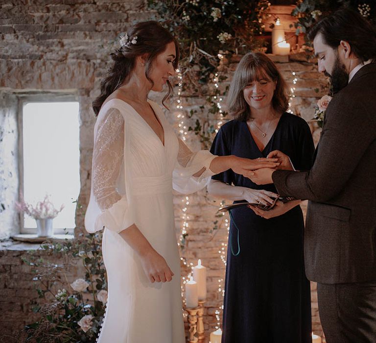 The bride and groom exchange rings for their rustic winter wedding with soft candlelight and fairy lights 