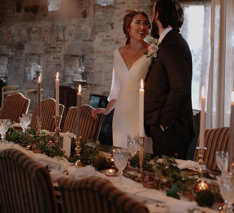 Banquet table with white and grey taper candles, foliage table runner, and crystal wine glasses with the bride and groom in black tie