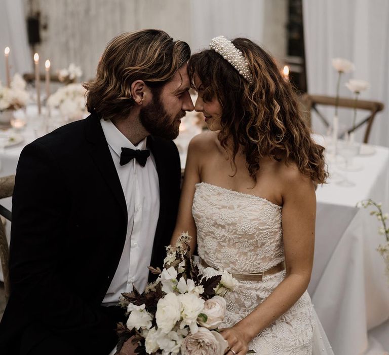 Groom in black tux and bride in sleeveless lace wedding dress with pearl details, pearl headband and silver belt embracing