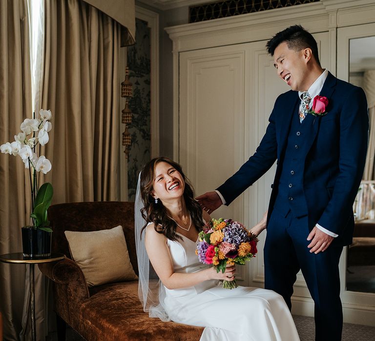 Bride holds brightly coloured floral bouquet and sits beside her groom in three-piece suit with pink rose buttonhole 