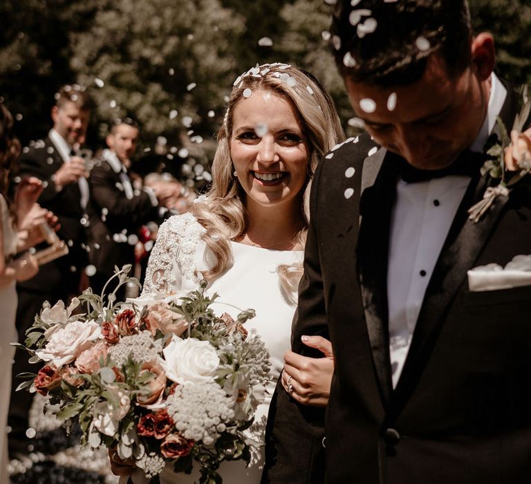 Groom in black tie leads the bride through the confetti with a pink and white bridal bouquet 