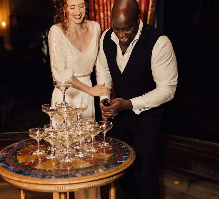 Groom in black waistcoat and suit trousers with bride in two piece outfit with sparkly long sleeve top and satin wide leg trousers topping up champagne tower 