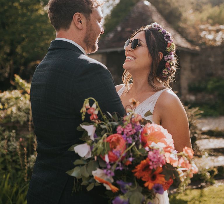 Bride wearing sunglasses and hugs her groom whilst holding colourful floral bouquet 