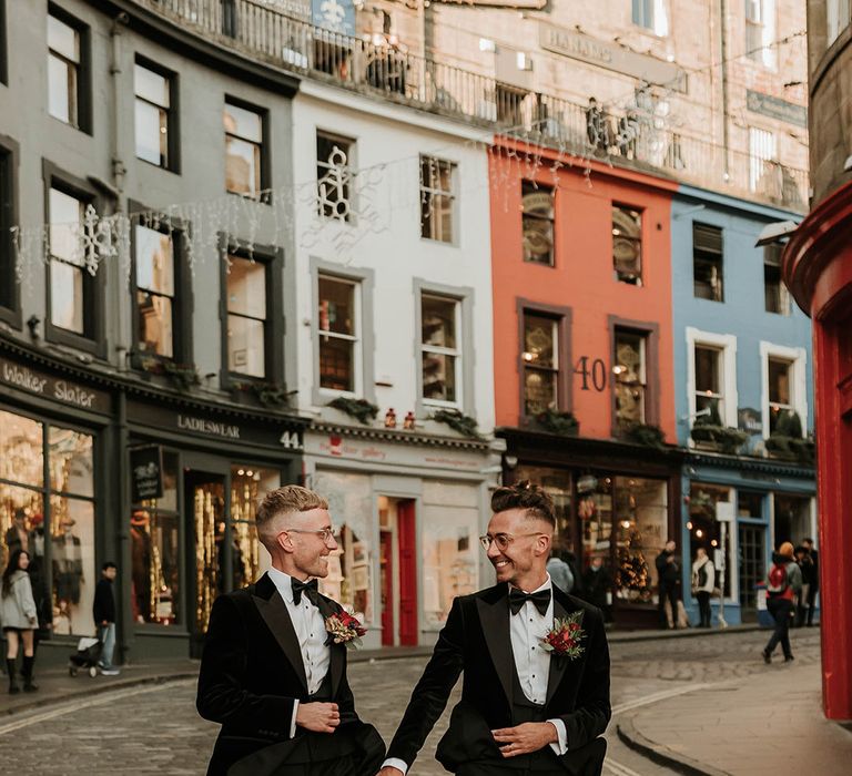 Grooms in matching black velvet tuxedos with black bowties and red rose and foliage boutonnieres walking the streets of Edinburgh city centre after stylish, contemporary wedding