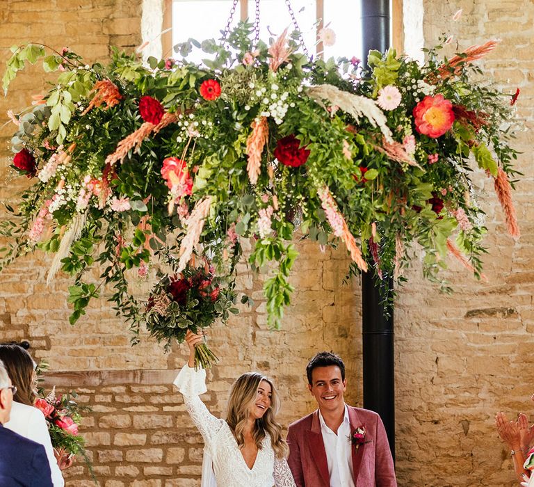 Bride holding up bouquet with red, pink and white garden roses, eucalyptus, foliage and dried flowers underneath suspended foliage wedding decor wearing  long sleeve lace boho wedding dress with tassels and groom in pink corduroy suit 