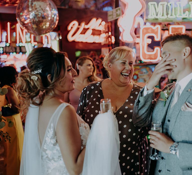 Bride in sleeveless plunge lace wedding dress and cathedral length embroidered veil laughing with wedding guests at Gods Own Junkyard 