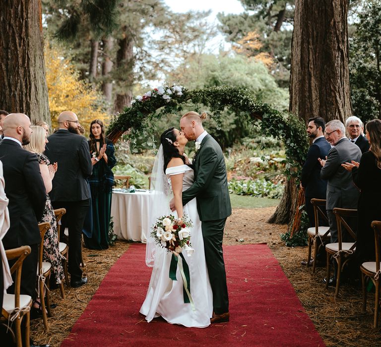 Bride holds white orchid bridal bouquet and she kisses her groom in green suit in front of floral archway at the Royal Botanic Garden Edinburgh