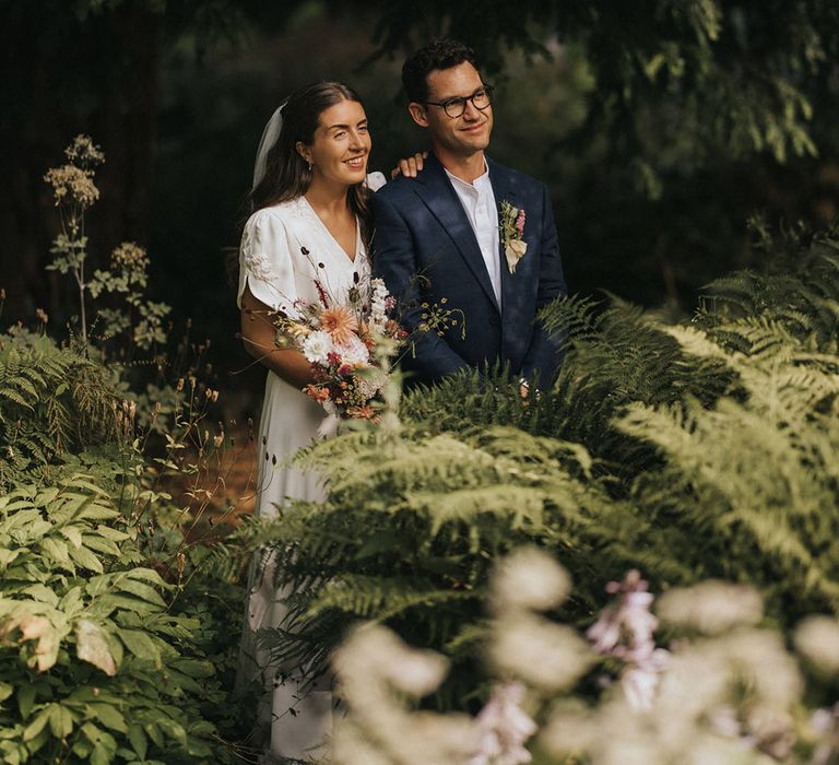 Groom in navy suit with white shirt with the bride in embroidered wedding dress with rustic wedding flowers 